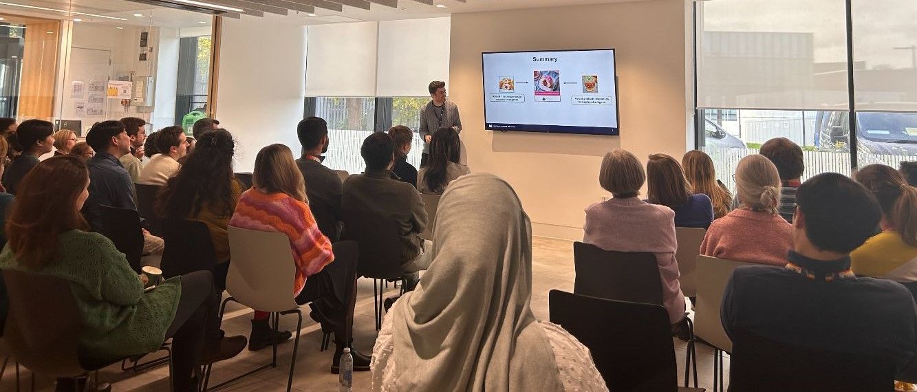 Speaker stands in front of a screen displaying their research data talking to a seated audience, who have their back to the camera.