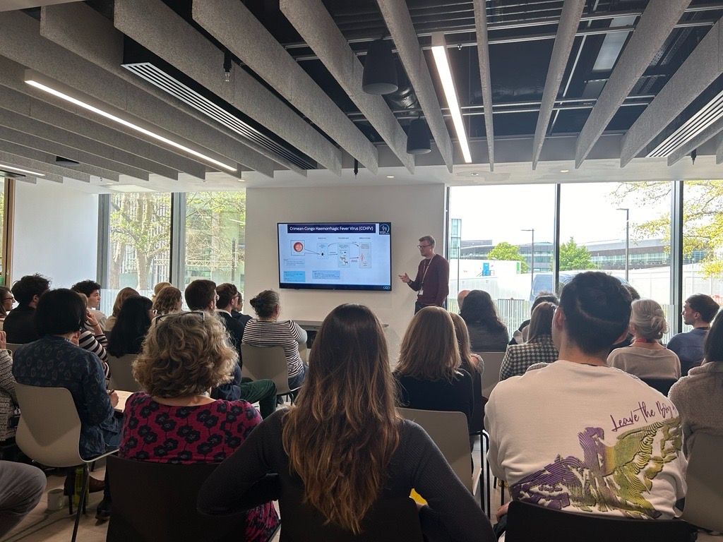 DPhil student speaker stands in front of a screen displaying their research data talking to a seated audience, who have their back to the camera.