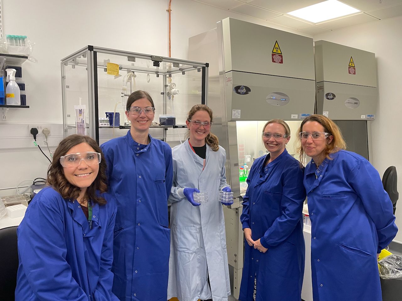 Three school teachers wearing blue lab coats take a break from an experiment in the lab and stand in a line with two researchers to smile at the camera.