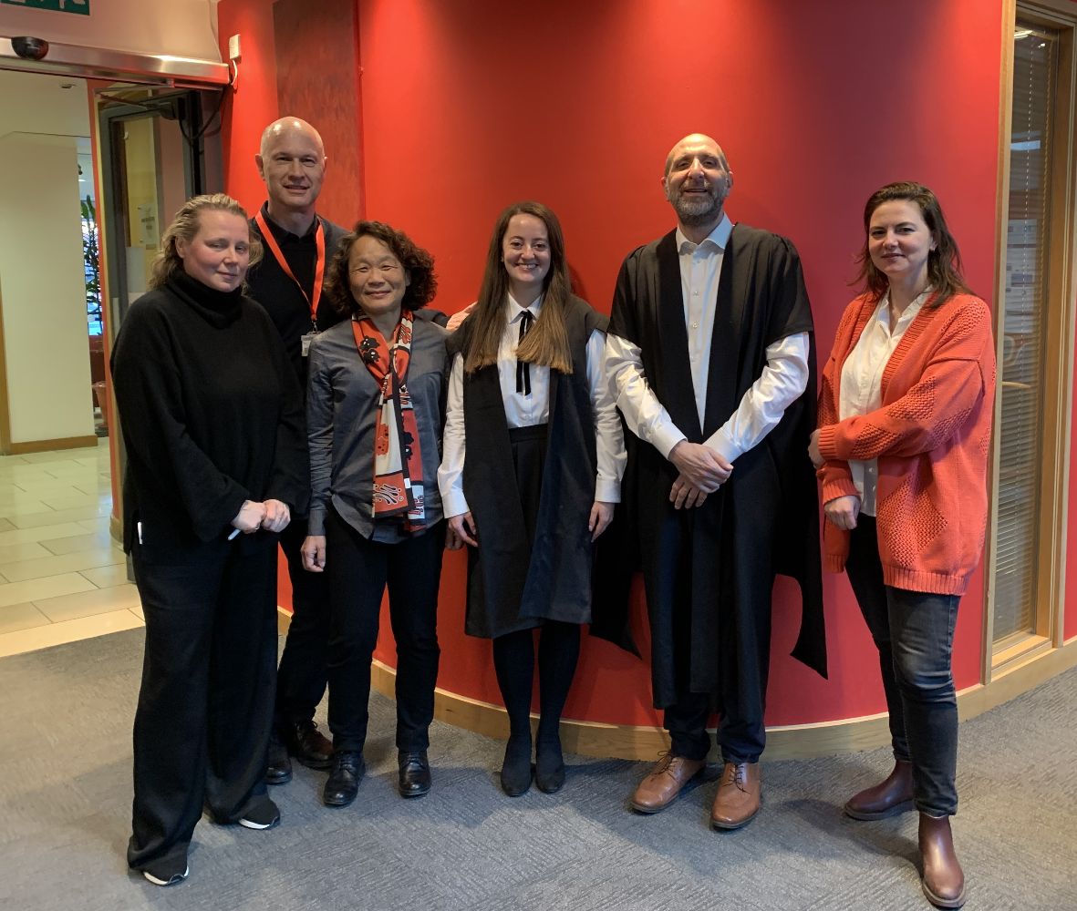 Six people stand shoulder-to-shoulder facing the camera in front of a red wall.
