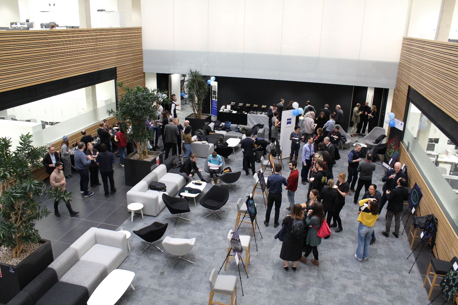Crowd of people mingling in a large, open atrium with enlarged microscopy images displayed on easels, viewed from an elevated perspective