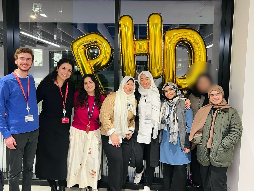 Eight people stand in a line infront of helium filled balloons spelling PHD